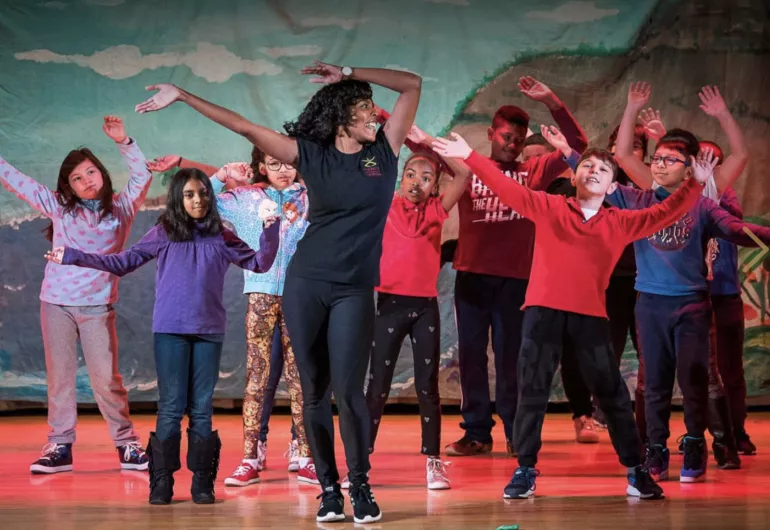 A teacher wearing black pants and t-shirt stands in front of a group of children, all with their arms up over their heads in dance poses.