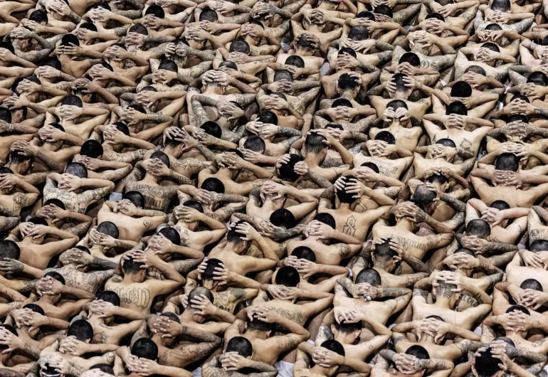 A view of hundreds of El Salvadorian prisoners sitting with their hands clasped behind their heads, exposing bare shoulders.