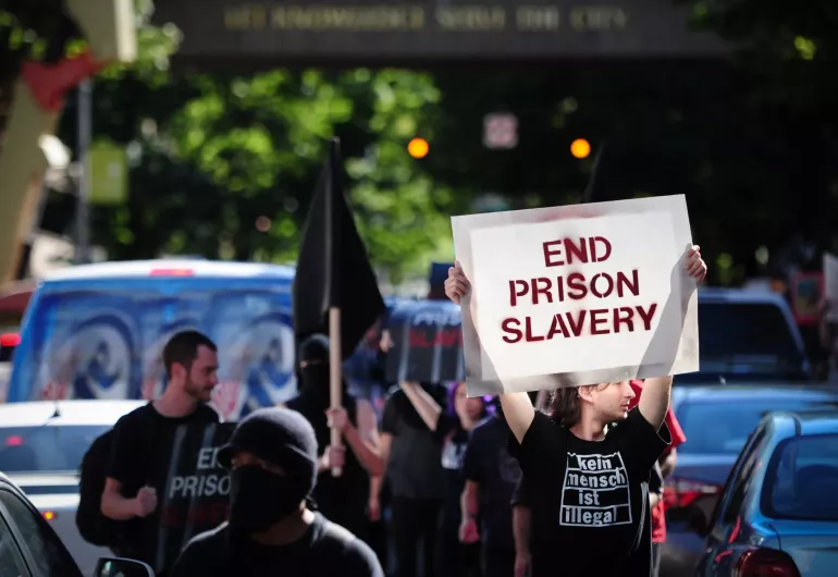 Crowd marches down a street holding a sign saying "End Prison Salvery"