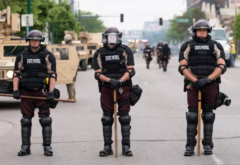 Three Minnesota State Patrol officers in riot gear guarding a street with armored vehicles behind them