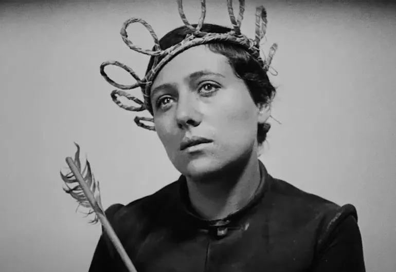 A black-and-white close-up of a woman with a rope crown and holding a feathered arrow, looking contemplatively upward.