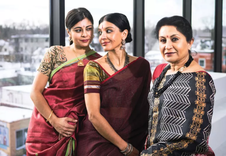 Ashwini, Aparna, and Ranee Ramaswamy dressed in red, burgundy and black and white patterned saris, stand close to one another.
