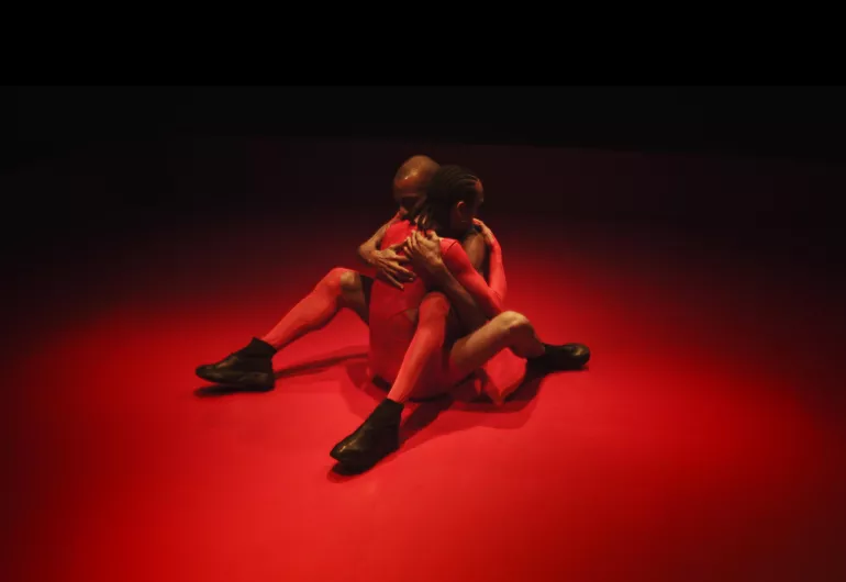 Dancers Pitts and Fredericks perform a sitting embrace. The image is linked to the Touch of RED teaser video on YouTube.