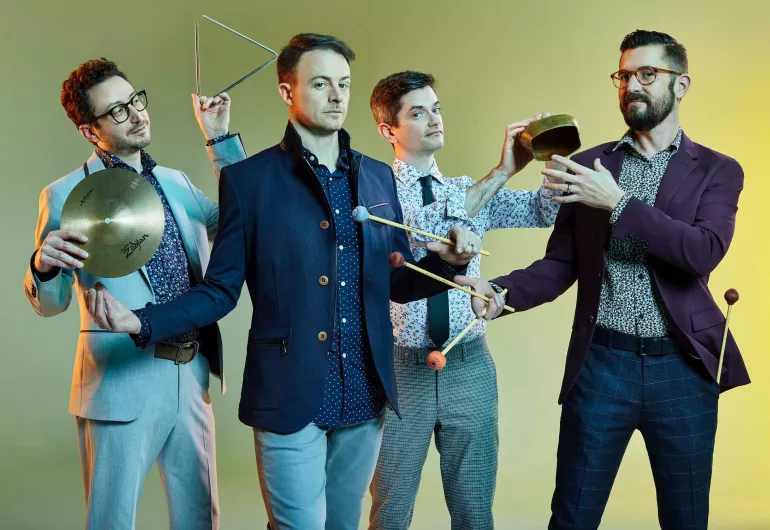 Four men, who are members of Third Coast Percussion, are wearing colorful jackets and button-up shirts and holding various percussion instruments.