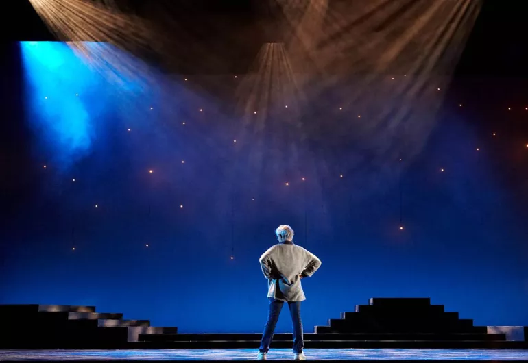 Twyla Tharp stands on stage with her back turned under spotlights.