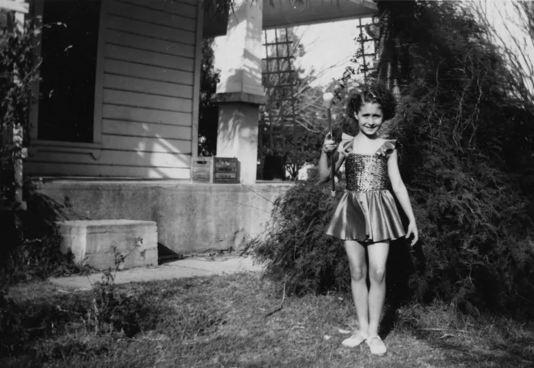 Twyla Tharp as a young, smiling child wearing a dress holds a baton outside a house.