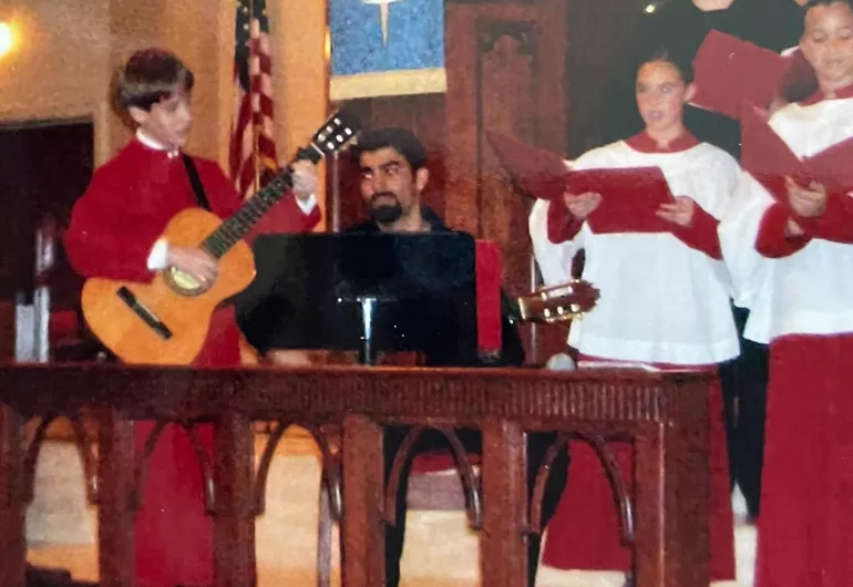 A young boy in a choral robe plays guitar alongside his instructor and church choir.