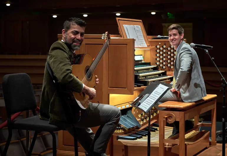 A man with a guitar smiles next to a smiling man sitting at an organ.