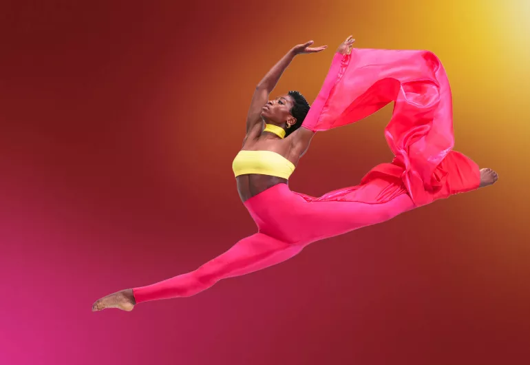 A dancer in a colorful costume, mid-split leap. Image is linked to a YouTube video.