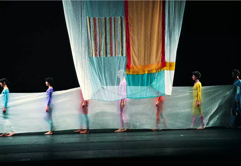 Six dancers in colorful outfits stand behind sheer fabric under fabric hanging down from above.