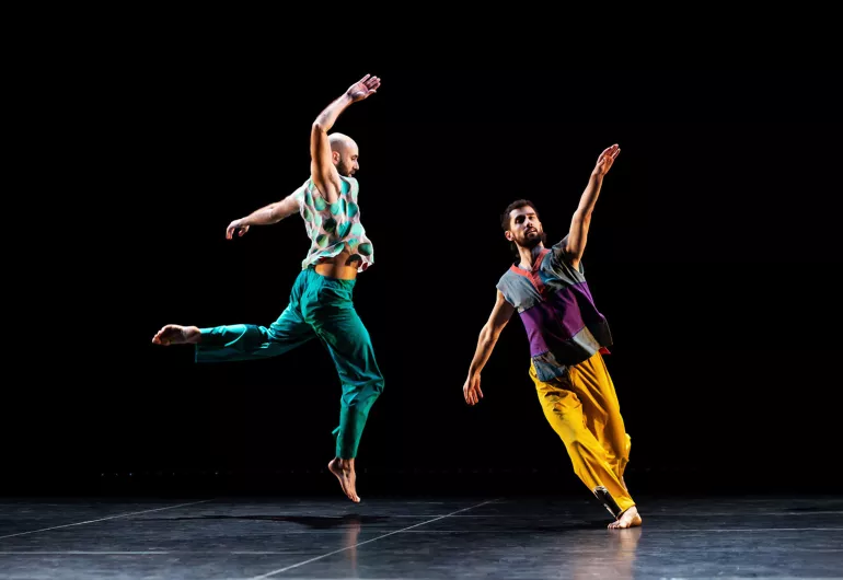 Two dancers from the Trisha Brown Dance Company performing on a dark stage, wearing colorful outfits.