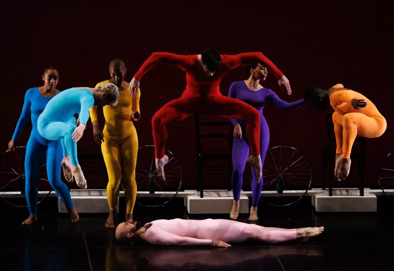 Dancers in different colored unitards jump with legs and arms out, one lies on the floor in the middle.