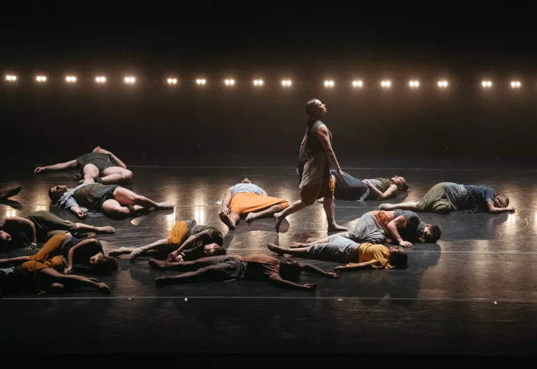One dancer steps forward while several dancers lay collapsed on the stage around her.