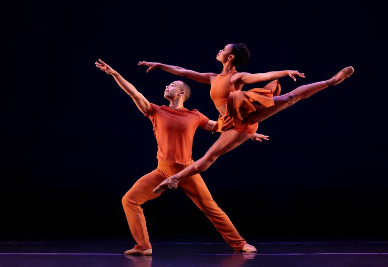 Two dancer in orange dance against a black background.