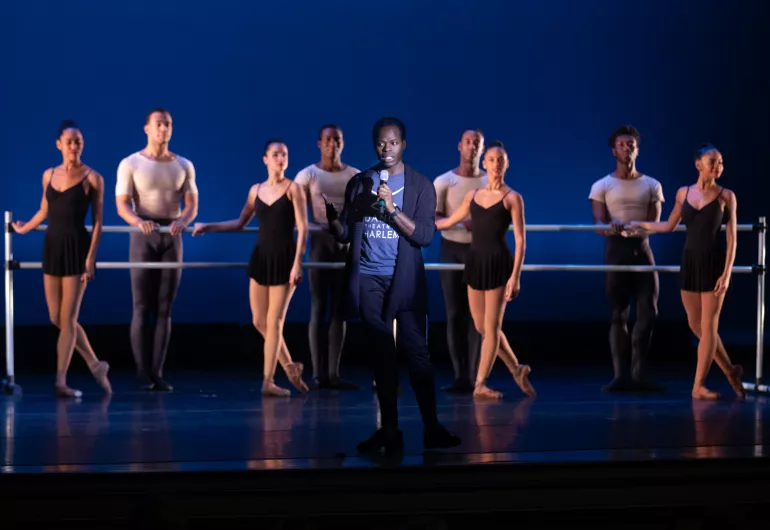 A man holds a microphone on stage with a line of dancers behind him.