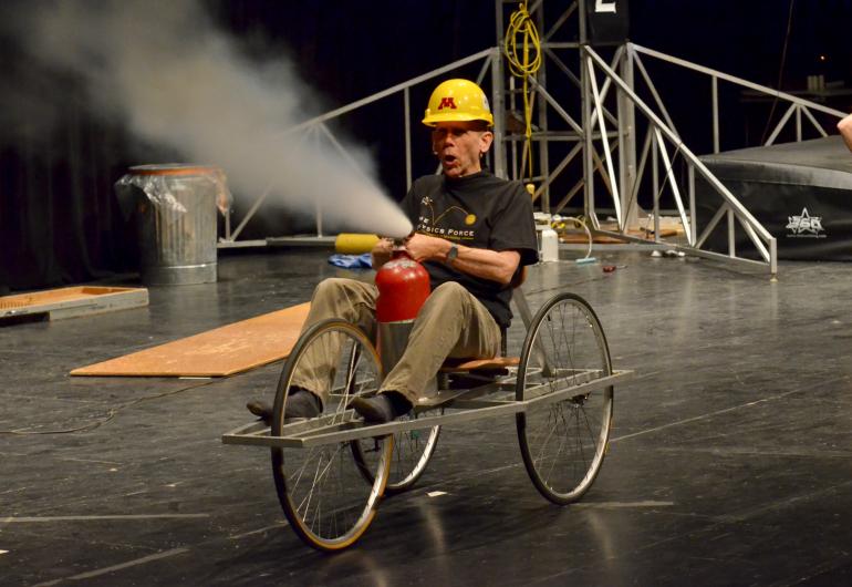 A man sits on a tricycle while spraying a fire extinguisher. 