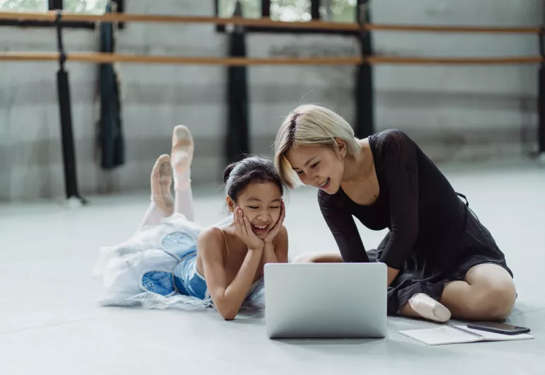 A young ballet dancer on the floor looking at a laptop with a smiling adult