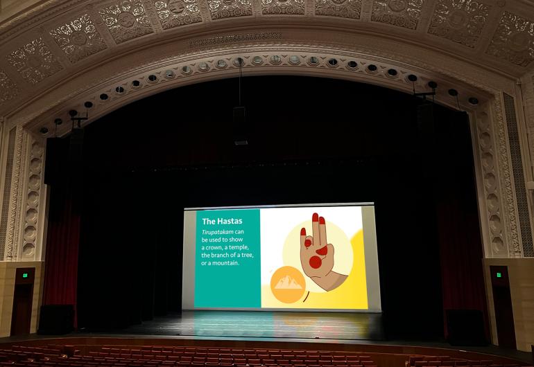  A large auditorium that has detailed molding outlining the stage, shows a projected screen with an image of the hand symbol for The Hastas and a small orange circle with a graphic of mountains in it. The screen has text on the left side that reas, “Tirupatakam can be used to show a crown, a temple, the branch of a tree, or a mountain.”