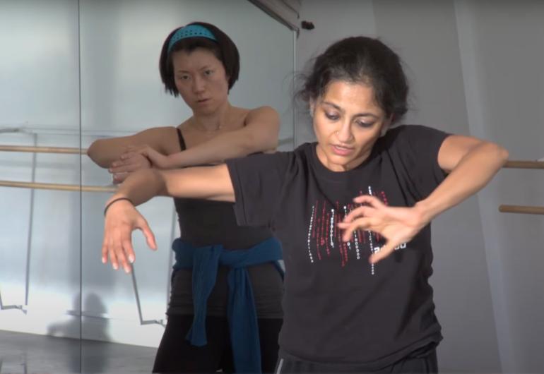 Two females dancing in a rehearsal. The female in the foreground is wearing a black t-shirt.