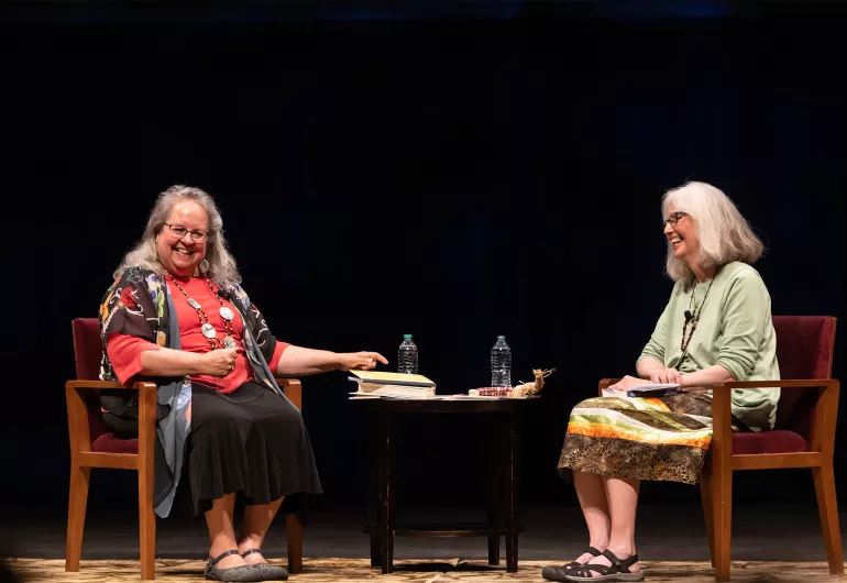 Two women with medium length grey hair, sit in wooden chairs facing eachother with a table between them, on a stage with a black backdrop. 