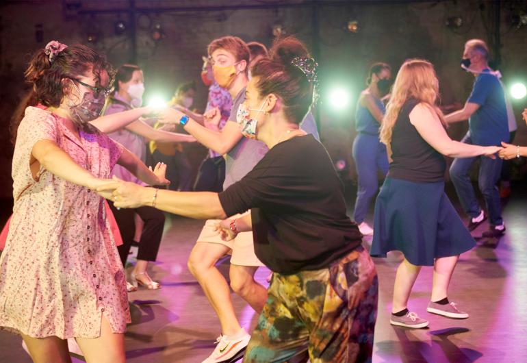 A class of participants wearing masks learning to swing dance on the Northrop stage. In the foreground are two women holding hands and practicing the movement.