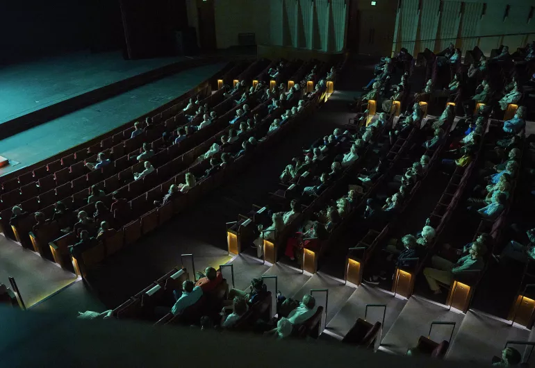 Audience members sit in theater seats watching the stage. Everyone is washed in a teal light emanating from the performance onstage.