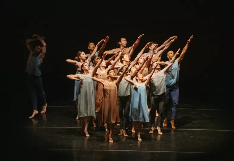 A group of dancers stands illuminated on a black stage, pointing their right arms up on a diagonal and looking in the same direction. Another dancer stands to the left with both arms above their head in the dark.