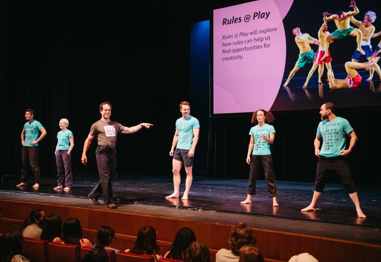 Five Pilobolus dancers wearing light blue-green t-shirts and black bottoms stand on-stage smiling, while a dancer wearing a black t-shirt and black pants walks in front of them holding one arm out pointing. A pink projection behind them says, Rules at Play. Caption: Matt Kent, artistic director of Pilobolus, leads an interactive discussion with the audience at the Student Matinee: Rules @ Play.