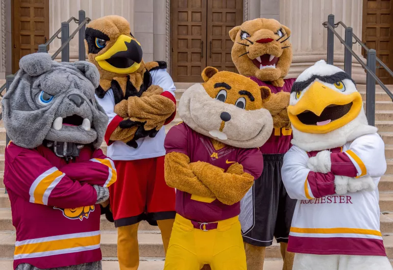 Five University of Minnesota mascots posing together with arms crossed on building steps.