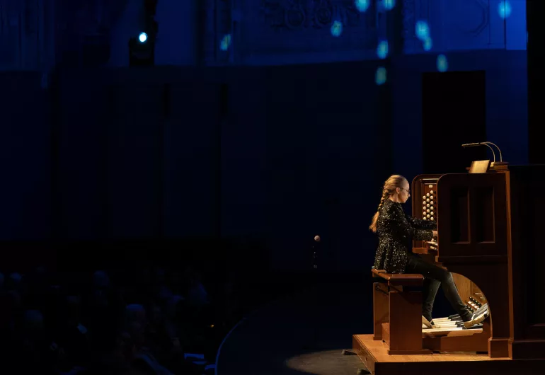 Anna Lapwood with blonde braided hair in a black sequin blazer looking down and playing a pipe organ. 