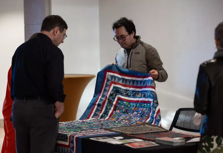 A man looks at a quilt that another man is showing at a display table.