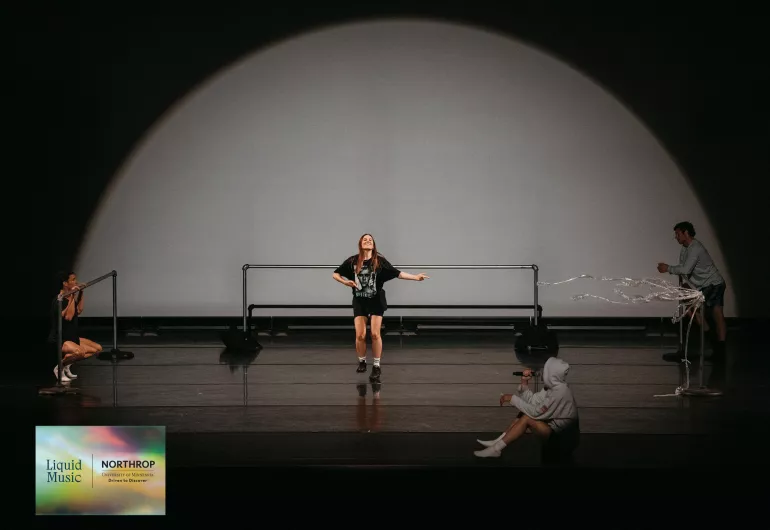 Performer tap dancing in a black shirt on the stage with 3 people surrounding her.