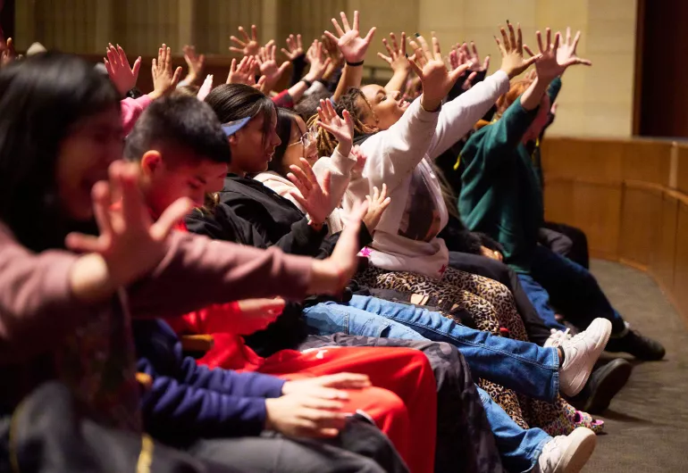 K-12 students wave their hands and reach forward in unison during a performance.