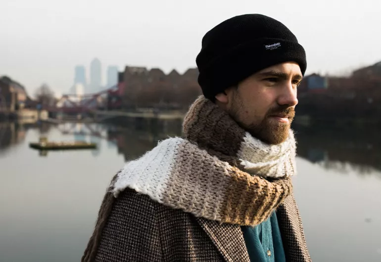 Kit Downes standing in front of a river wearing a scarf, jacket, and hat.