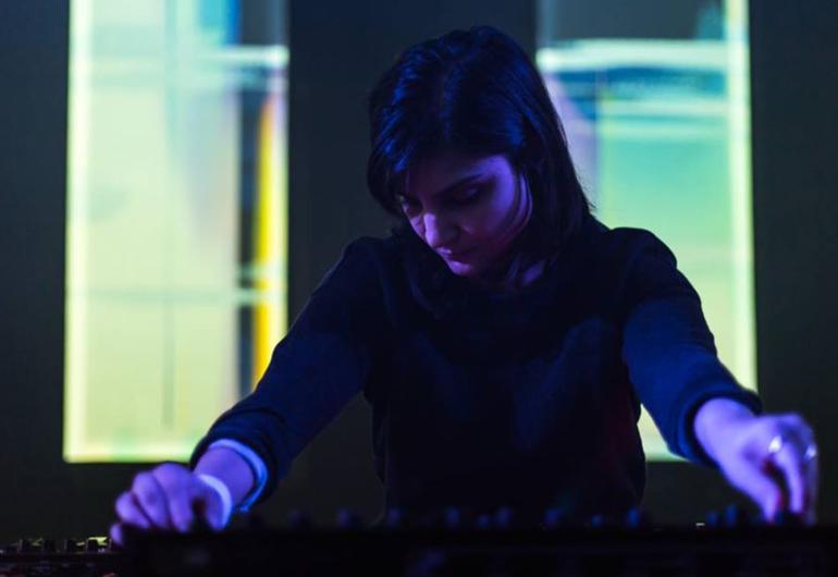 Sarah Davachi in dark clothes and purple light with light pastel colors behind her