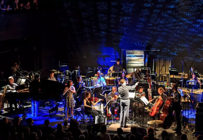 Alarm Will Sound, a small orchestra of about 16 musicians and a conductor, performs on stage.
