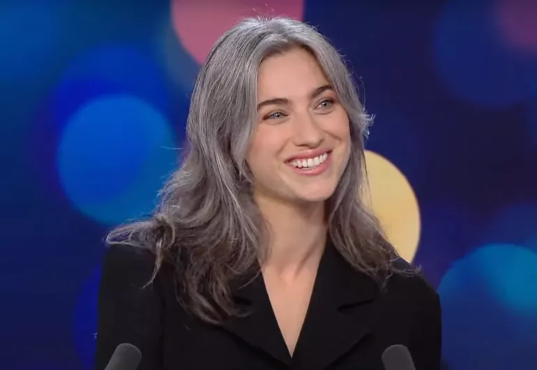 Kali Malone, a young woman with grey hair, wears a black blazer and smiles. Link to France 24 video - YouTube.