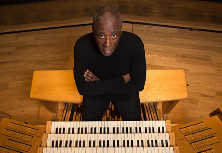 Wayne Marshall, a man wearing all black clothing, sits behind a four-level organ keyboard