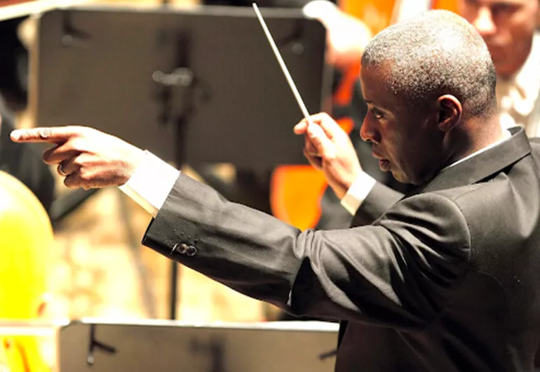 A sideview of Wayne Marshall wearing a dark suit with arms up and a conductor's baton in his right hand conducting an orchestra which is blurred in the background