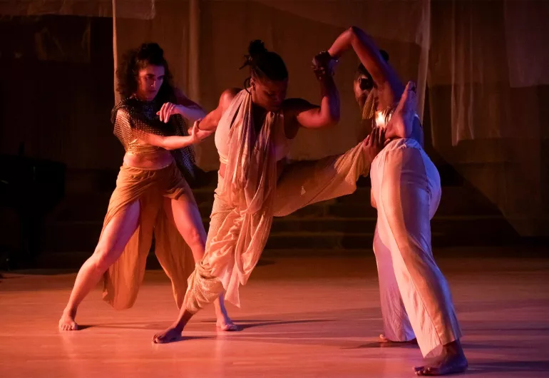 three dancers in white moving together on a stage with pinkish light around them