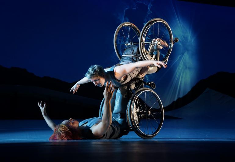 Laurel Lawson, a white woman, is flying in the air with arms spread wide, wheels spinning, and supported by Alice Sheppard. Alice, a light-skinned Black woman, is lifting from the ground below. They are making eye contact and smiling. 