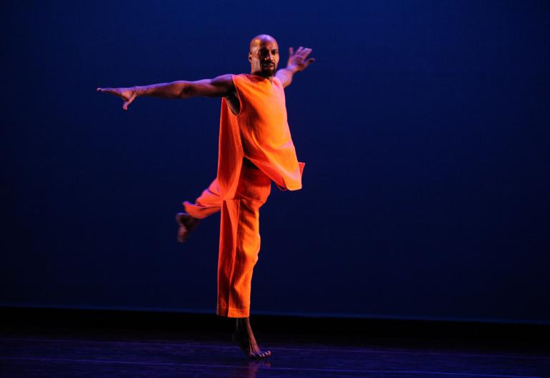 A male dancer dressed in bright orange performs against a purple background.