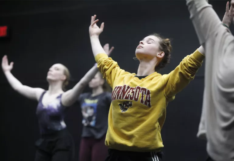 Young woman in gold UMN sweatshirt reaching up while dancing.