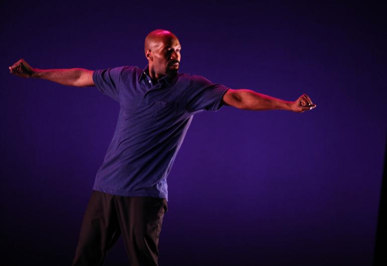 A male dancer in a purple shirt performs against a purple background.