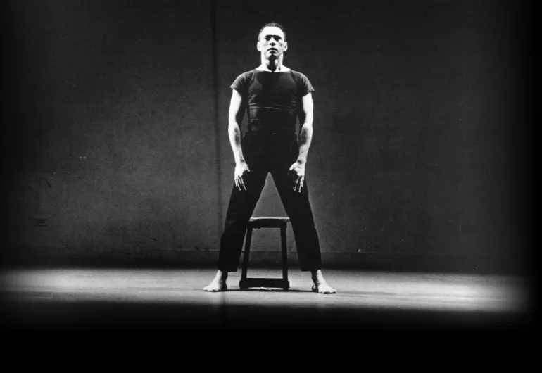 Victor Quijada stands firmly over a stool, legs apart and arms down, in spotlight against black background.