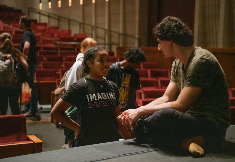 A young female student with brown skin and braided hair, wearing a black t-shirt with the word "Imagine" stands at the foot of a stage speaking to a man with light skin, brown curly hair, wearing  a camo t-shirt.