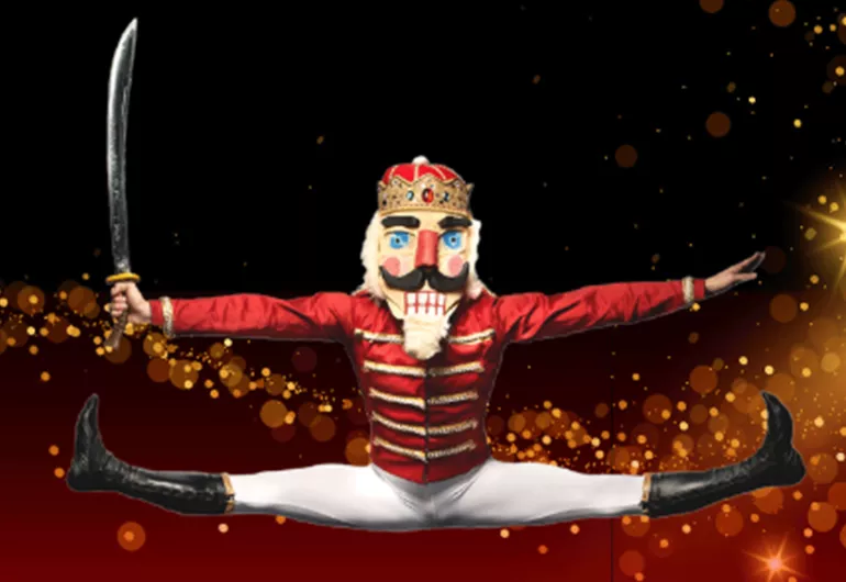 Nutcracker character in open jump splits, arms out to the side, holding a sword.