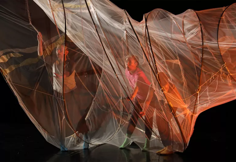 Performers interacting with a large, translucent fabric structure under dynamic stage lighting.
