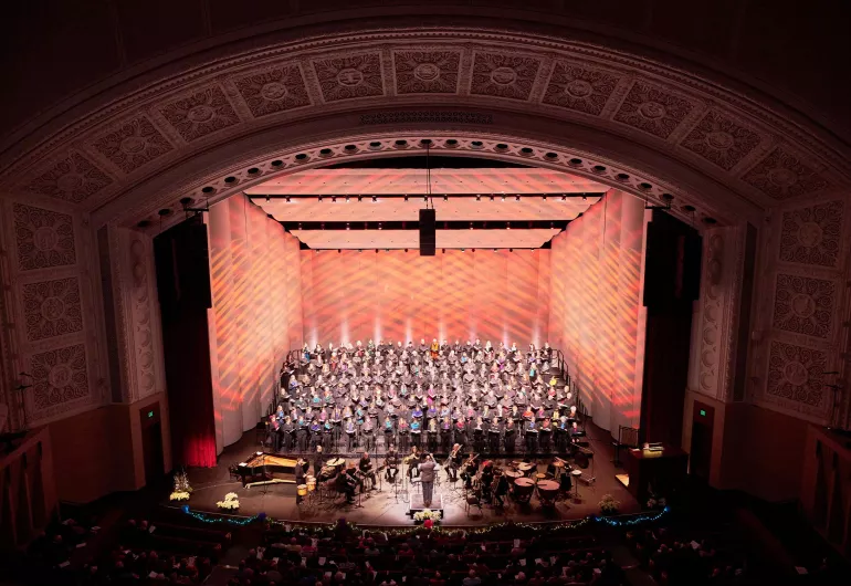 A choir and music ensemble perform on a stage in a grand theater with a high, ornate ceiling and warm red lighting.