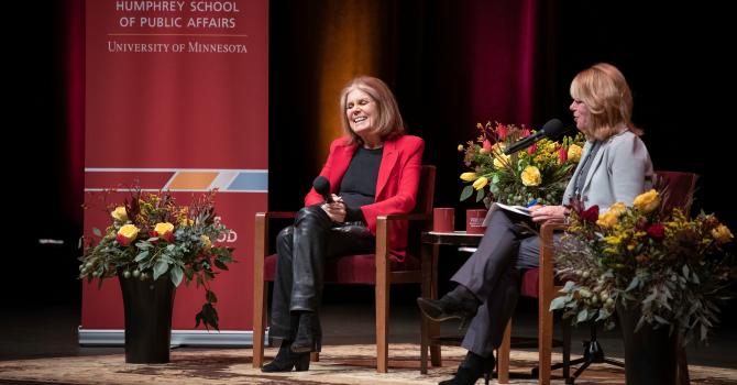 Gloria Steinem in a red jacket seated on stage next to a woman in a blue jacket